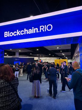 A photograph of the bustling entrance to the 'Blockchain.RIO' event, featuring a large blue illuminated sign and a crowd of attendees.
