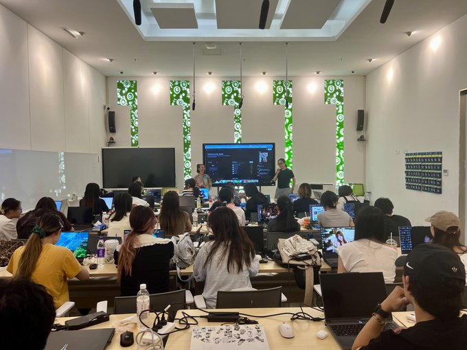 A well-lit classroom or conference room is filled with attendees working on laptops while two presenters stand at the front near large screens.