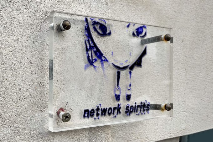 A clear acrylic sign mounted on a textured wall with metal standoffs, featuring blue graffiti-style artwork of a face and the text 'network spirits'.