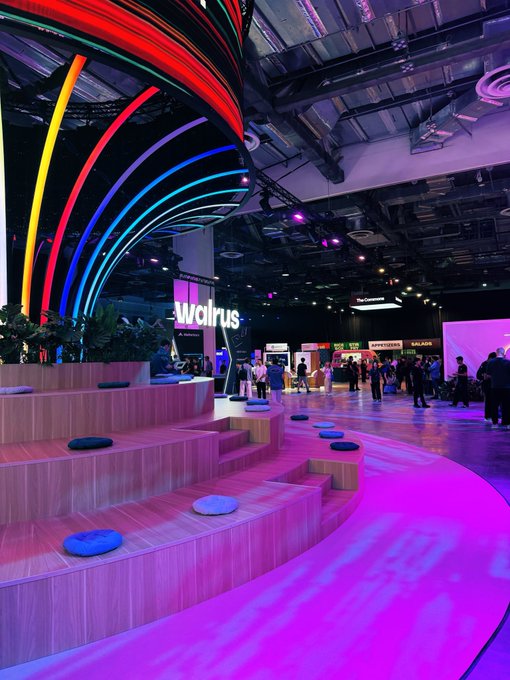 A vibrant indoor event space features a colorful neon arch installation in red, yellow, green, and blue, with a "walrus" sign visible and tiered wooden seating with circular cushions in the foreground, bathed in pink lighting.