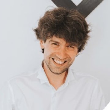 The image features a man wearing a white shirt, smiling at the camera with his eyes closed. The background of the photo shows a wall that has a black x on it. There are no other objects or people visible in the image, so the focus is solely on the man and the context provided by the post.