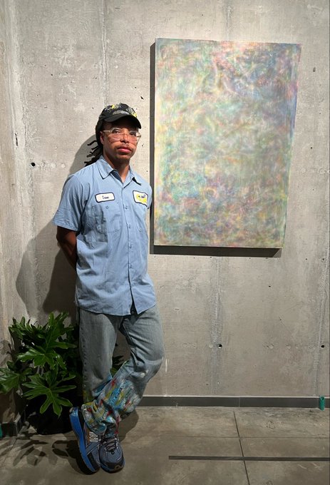 A man wearing a cap, glasses, and a blue work shirt stands next to a large abstract painting mounted on a concrete wall, with a green plant beside him.