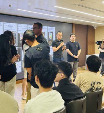 A photograph captures an indoor gathering where two men stand at the front of a room holding microphones and presenting to a seated audience, with academic posters displayed on the wall behind them.
