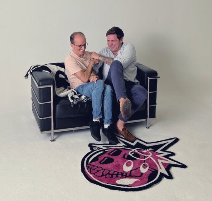 Two men laugh together while sitting on a black leather couch in front of a pink skull-shaped rug.