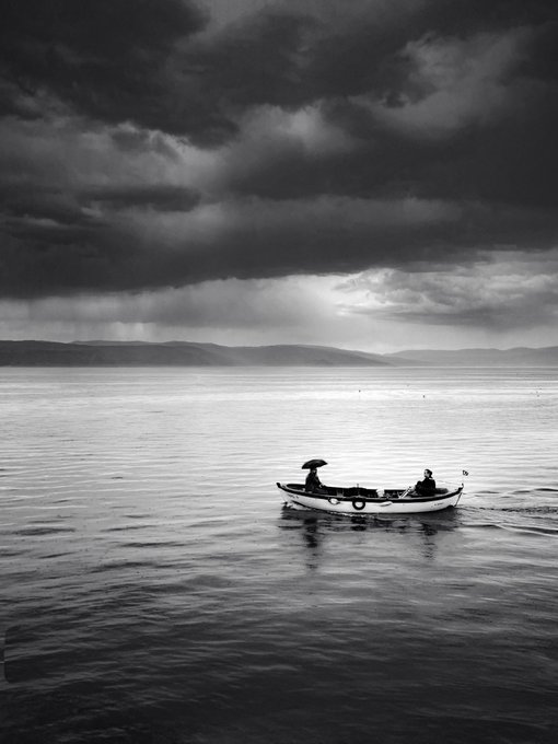 A black and white photograph of a small boat carrying two people with umbrellas across a calm body of water under a heavy, cloudy sky.