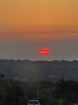 A sunset scene with an orange-red sky, a red sun setting behind a hazy horizon of trees, with the top of a vehicle visible at the bottom of the frame.