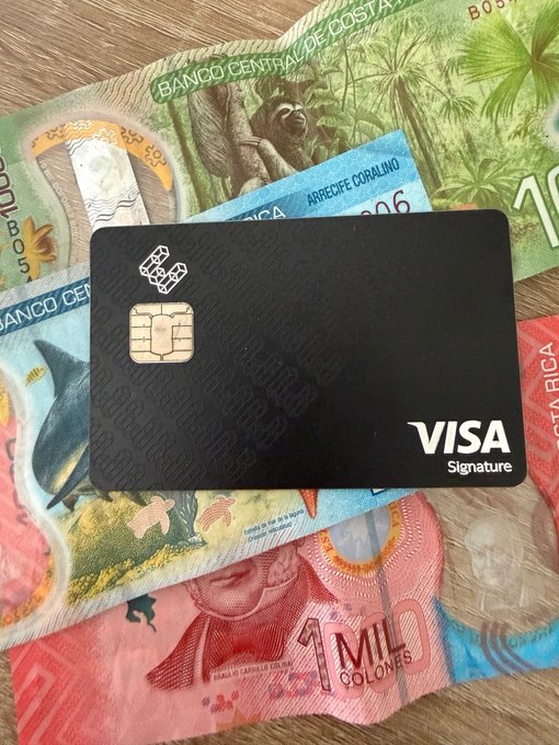 A black Visa Signature credit card resting on colorful Costa Rican colón banknotes from the Banco Central de Costa Rica, including a 1,000 colones bill featuring wildlife and a portrait.