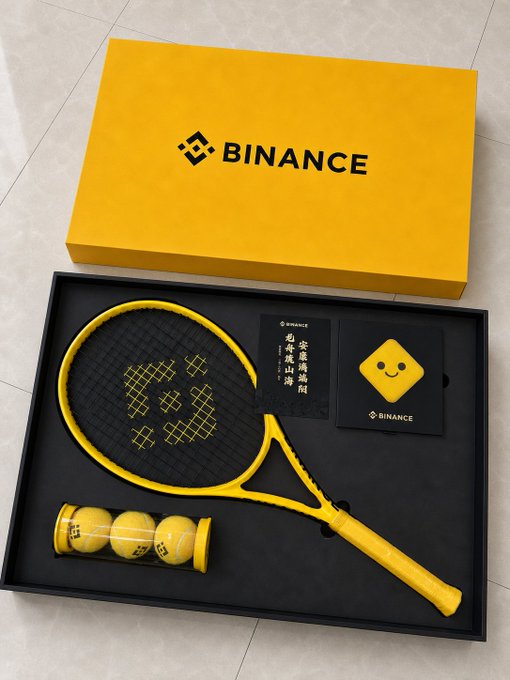 The image features a tennis racket, ball, and box with a yellow label that says "BINANCE" written on them. The tennis racket has a yellow handle and yellow frame, while the tennis balls are also yellow. The box contains more tennis balls as well as a small black and white square card with a smiley face drawn on it.