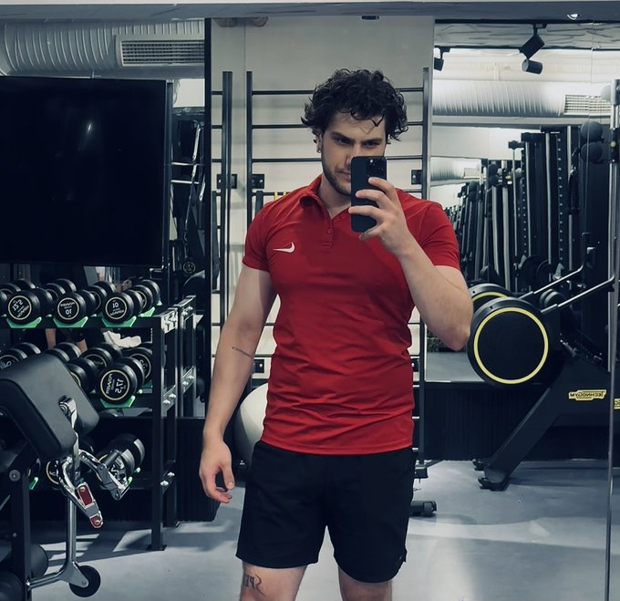 A gym mirror selfie of a man in a red Nike shirt and black shorts holding a phone, standing in a gym with dumbbells and exercise equipment visible.