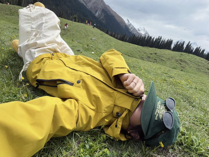 A person in a yellow raincoat lies on their back in a grassy mountain meadow, wearing a green cap with sunglasses on top, with white pants pulled up toward their head.