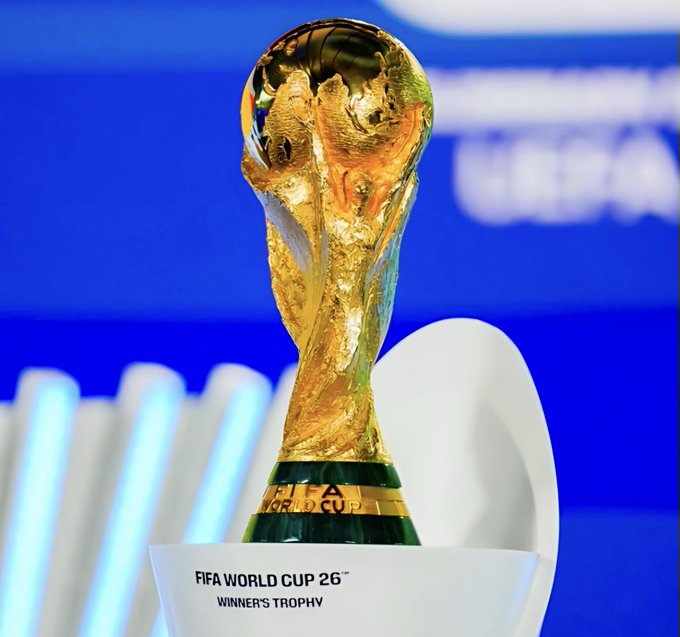 The image features a large, golden trophy that resembles the FIFA World Cup trophy. It is placed inside an open toilet bowl with a white lid. The text "FIFA World Cup 26 Winner's Trophy" is visible next to the trophy. The background of the image shows a blue wall and some books stacked on top of each other.