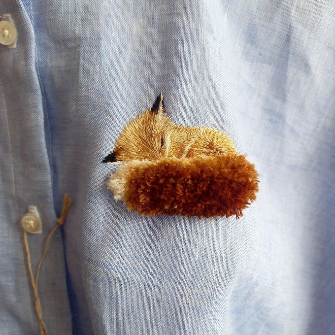 A needle-felted brooch depicting a sleeping fox with a fluffy reddish-brown tail is pinned to a light blue button-up shirt near the collar.
