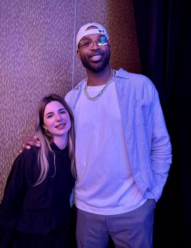 Two people pose together in a warmly lit room, with a smiling woman in a black top standing next to a tall man wearing a white cap, glasses, and an unbuttoned blue striped shirt over a white tee. The image likely accompanies a discussion about Tristan Thompson's continued involvement in crypto despite many mainstream celebrities leaving after the NFT boom faded.