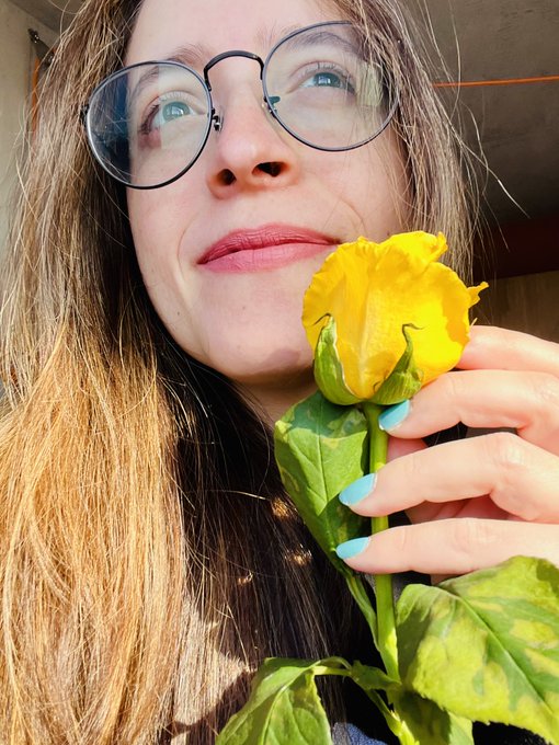 A close-up selfie of a smiling woman with glasses holding a yellow rose, showing her turquoise painted nails.