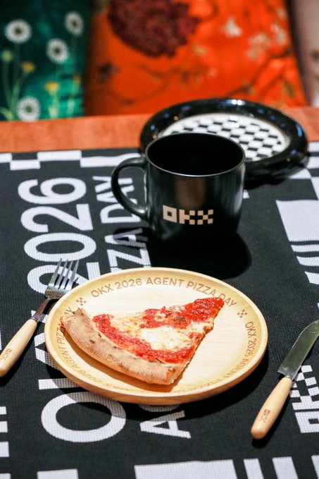 The image features a slice of pizza served on a wooden plate, accompanied by a fork and knife. The pizza has been cut into eight equal slices, making it easy for someone to enjoy their meal. In addition to the pizza, there are two cups in the scene - one black coffee cup and another white mug with a logo. A person is also visible in the background, but they are not the main focus of the image. The post that accompanies this image reads "GM 用 OKX 餐具吃早餐", which seems to be related to the pizza or the dining experience as a whole.