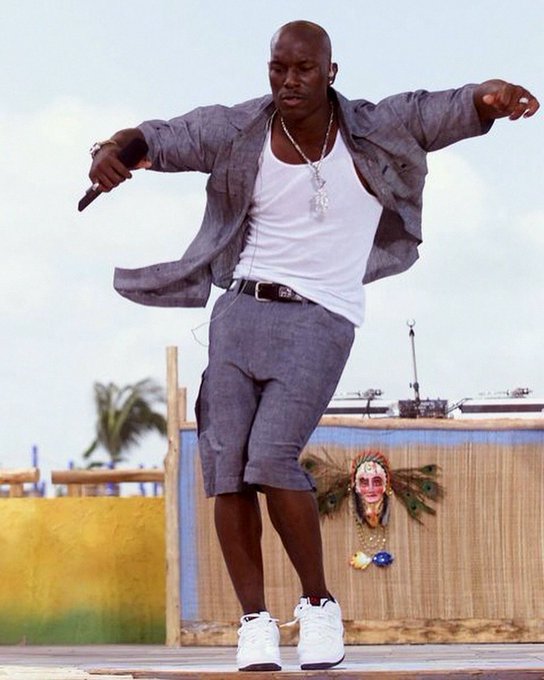 A man wearing a white tank top, grey open shirt, grey shorts, and a cross necklace performs a dance move on an outdoor stage, holding a microphone while wearing white sneakers.