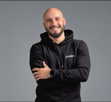 A smiling bald man with a beard stands with arms crossed wearing a black 'ZAMA' branded hoodie.