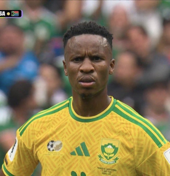 A South Africa national football team player wears the yellow and green jersey with the South Africa badge and Adidas logo, with a blurred crowd in the background.