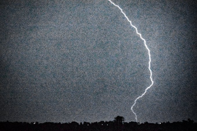 A nighttime photograph captures a bright, jagged bolt of lightning striking down toward a dark silhouette of a cityscape or town on the horizon.
