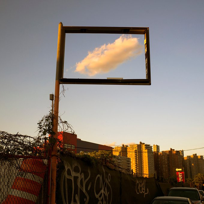An empty billboard frame against a blue sky shows a single golden-illuminated cloud passing through it, with a city skyline and a graffiti-covered wall visible below.