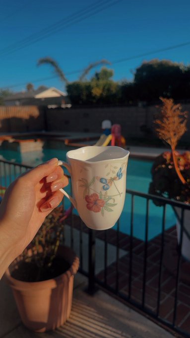 A hand holds a floral ceramic mug in the foreground while enjoying a sunny weekend by a backyard pool with palm trees and a playset in the background, capturing a relaxed morning vibe that pairs perfectly with the "Happy weekend $XRP Fam! What plans?" post.