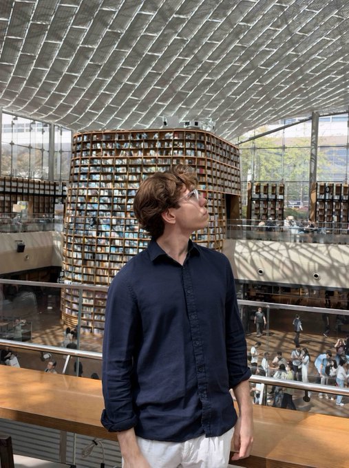 A young man in a navy blue shirt stands in front of a striking cylindrical library structure with bookshelves, looking upward at the architecture.