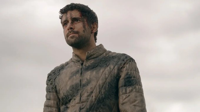 A man with dark curly hair and a beard, wearing a dirty and battle-worn medieval-style tunic, looks upward against a cloudy sky background.