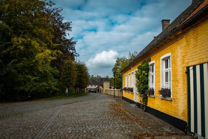 A cobblestone street lined with a yellow brick building featuring white-trimmed windows and flower boxes, with autumn trees in the background.