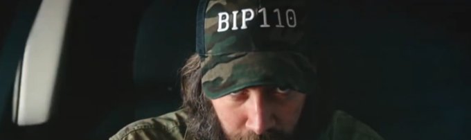 A cinematic still showing a bearded man wearing a camouflage cap with 'BIP110' written on it, appearing to sit inside a vehicle with a somber expression.