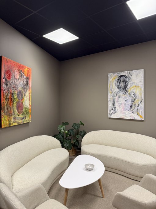 A modern lounge room with cream-colored curved sofas arranged around a white oval coffee table, set against dark walls adorned with two abstract paintings and a potted plant, under a black ceiling with square recessed lights.