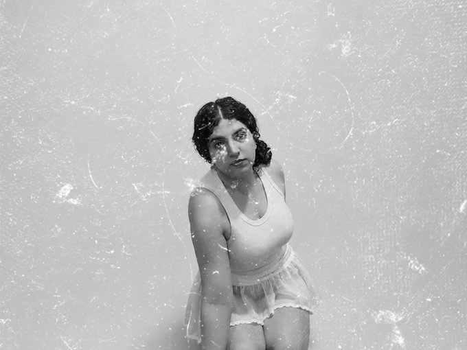 A black-and-white vintage-style photograph shows a woman with short dark hair wearing a white sleeveless top and skirt, gazing upward against a distressed, textured background—its raw, exposed aesthetic mirroring the caption's themes of vulnerability and shedding "Bare" on 11/11.
