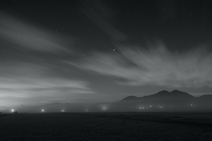 A black and white landscape photograph showing wispy clouds sweeping across a night sky over a dark mountain range, with scattered ground lights illuminating the horizon.