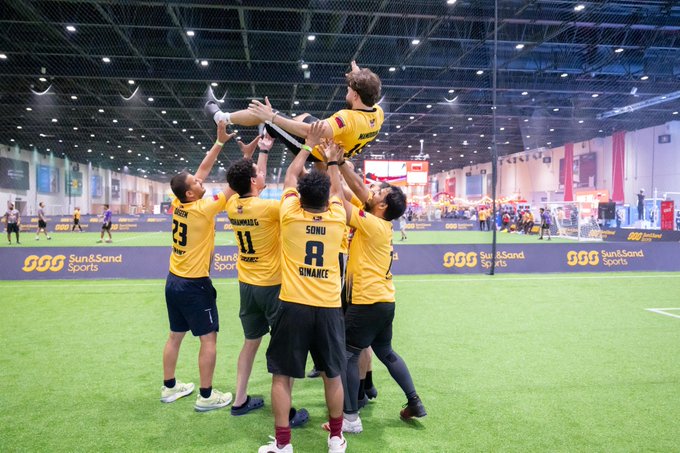 A group of players in yellow "Sun & Sand Sports" jerseys, including numbers 3, 8, and one named "Sons," hoist a teammate high into the air on an indoor turf field during a celebration. The lifting embrace playfully echoes the caption's playful claim of "touching grass," as the team takes a moment of triumph on the green playing surface.