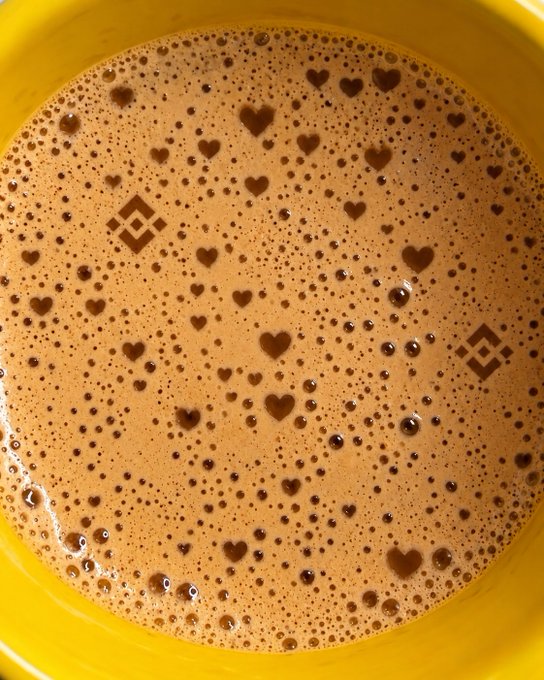 A close-up of a yellow cup filled with frothy brown beverage, possibly coffee or hot chocolate, with a Binance logo visible through the bubbles and heart-shaped patterns in the foam.