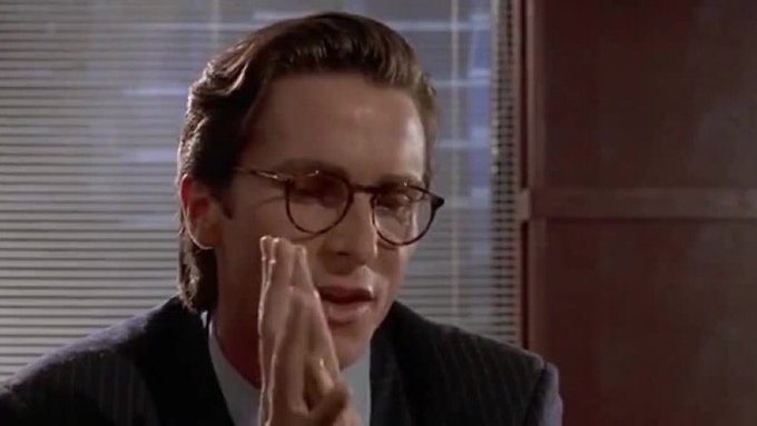 A man with slicked-back hair and glasses presses his hands together in a prayer-like gesture, wearing a pinstripe suit in what appears to be an office setting with blinds behind him.
