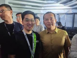 Two men posing together for a photo at an indoor event, with the man on the right wearing glasses and the man on the left wearing a lanyard with 'HKG' visible.
