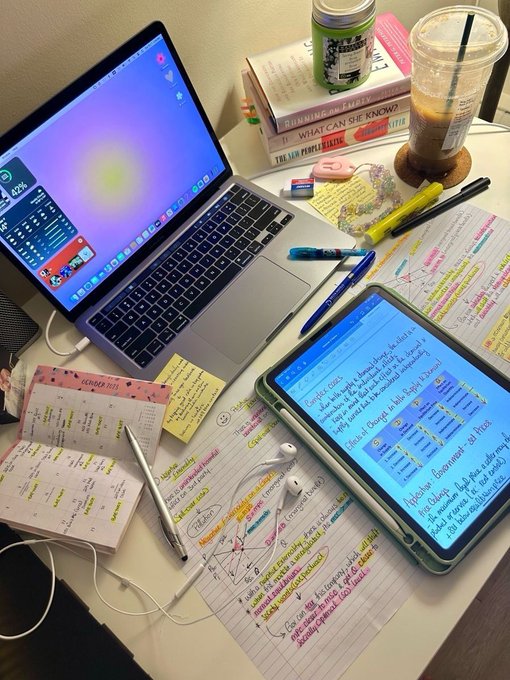 A cluttered study desk with a MacBook, iPad showing Google Slides about 'Climate Change & Sustainability', handwritten study notes, pens, sticky notes, earphones, and an iced coffee drink.
