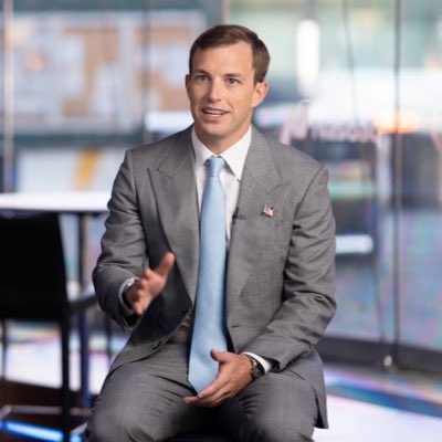 A professional photograph of Jared Kushner wearing a gray suit and light blue tie, gesturing with his hand while seated in a modern interior.