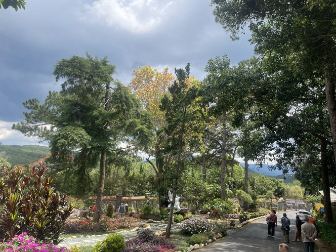 A scenic park view featuring lush green trees, flower beds, and mountains in the background under a partly cloudy sky, with visitors walking along a paved path.