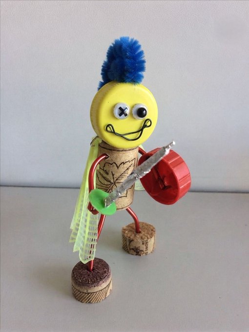 A whimsical handmade craft figure named "Bopper" stands on cork feet, featuring a yellow smiling face topped with a blue pom-pom, a wine cork body with a leaf stamp, and holding a bottle cork sword with a red bottle cap shield, decorated with a green plastic sword. This quirky upcycled creation represents the quirky, collectible NFT character set to launch on OpenSea via Robinhood, with the first 100 wallets earning the exclusive opportunity to own one.