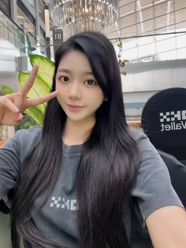 A selfie of a young woman with long dark hair wearing a grey t-shirt, making a peace sign with her hand in what appears to be a modern building interior.