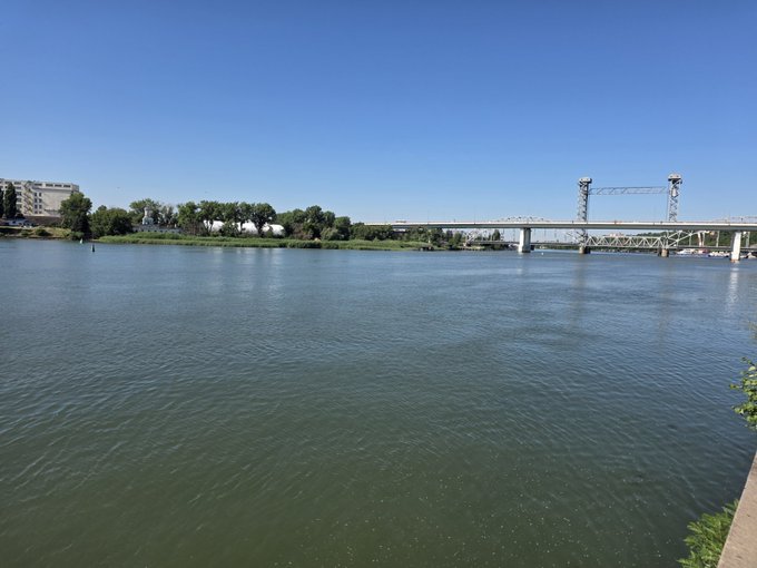 A scenic view of a river with a bridge and lift span in the background under a clear blue sky, with trees lining the far bank.