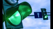 The image features a traffic light with its green light illuminated, indicating that vehicles can proceed through the intersection. Below the traffic light, there's a blue sign displaying an arrow pointing upwards, which suggests that drivers should look up from their current position to follow the direction of travel indicated by the arrow.