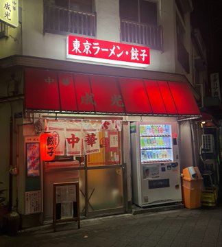 A nighttime photo of a Japanese storefront with a red awning and a sign reading '東京ラーメン・餃子' (Tokyo Ramen & Gyoza), featuring a vending machine and a lantern.