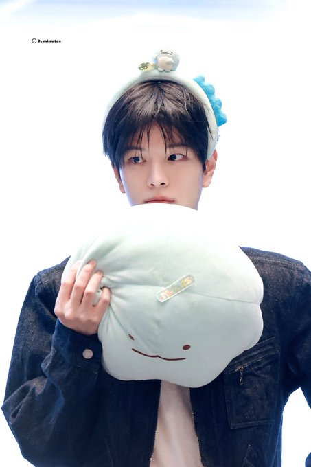 A young man wearing a denim jacket and a fluffy headband with cartoon characters, holding a smiling cloud-shaped plushie that covers the lower half of his face.