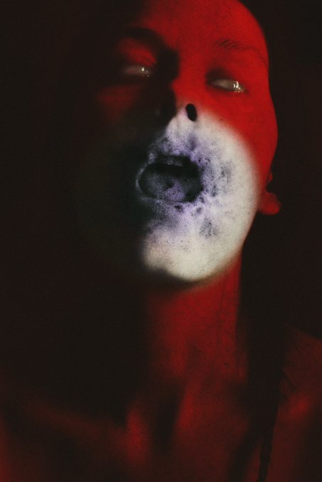 A person is bathed in red light with white makeup around their mouth, their head tilted back and eyes looking upward in a dramatic pose.