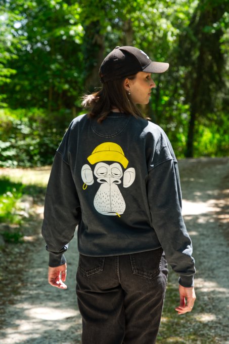 The image features a woman walking down a path, wearing a gray sweatshirt with a white graphic of a monkey on it. The backdrop includes a lush forest setting that surrounds her as she moves through the trail. There are no discernible texts or other objects in the image, and the woman is positioned near the center of the frame, indicating that she is at the forefront of the scene. The overall composition emphasizes the connection between art, fashion, and nature.