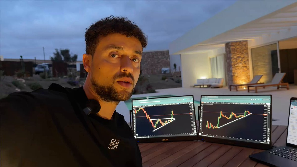 The image features a man sitting at a table with two computer monitors, one of which displays a stock chart or graph. The man appears to be wearing a black shirt and has a beard. There are also some chairs around the table where he is seated. In the background, there is a house visible through the window. The caption on the image reads "XRP: Today!!! (BEWARE)". This suggests that the man might be discussing or analyzing cryptocurrency trends in the context of the stock market.