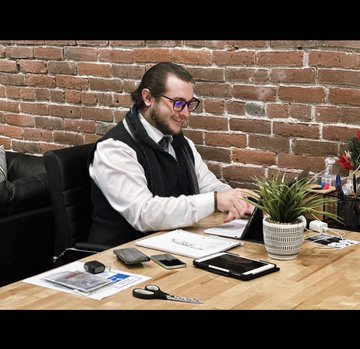 The image shows a man sitting at a wooden desk with his laptop open in front of him. He has a potted plant next to him and various office supplies such as pens, papers, and scissors scattered around the desk. The background features a brick wall, adding an urban touch to the scene. The post includes text that reads "Powerful message in Shibo's space this morning ... Reflected on how we climbed corporate America, hit CEO level, built a biz for my significant other - gutters to penthouse. Ego crept in and you start thinking you're too good for the 'normal' work". The man appears to be focused on his work while surrounded by these items.