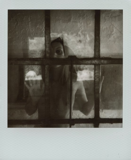 A vintage-style Polaroid photograph showing a person with dark hair and pale skin looking through a multi-paned window, holding a hand up to the glass in a dim, sepia-toned setting.